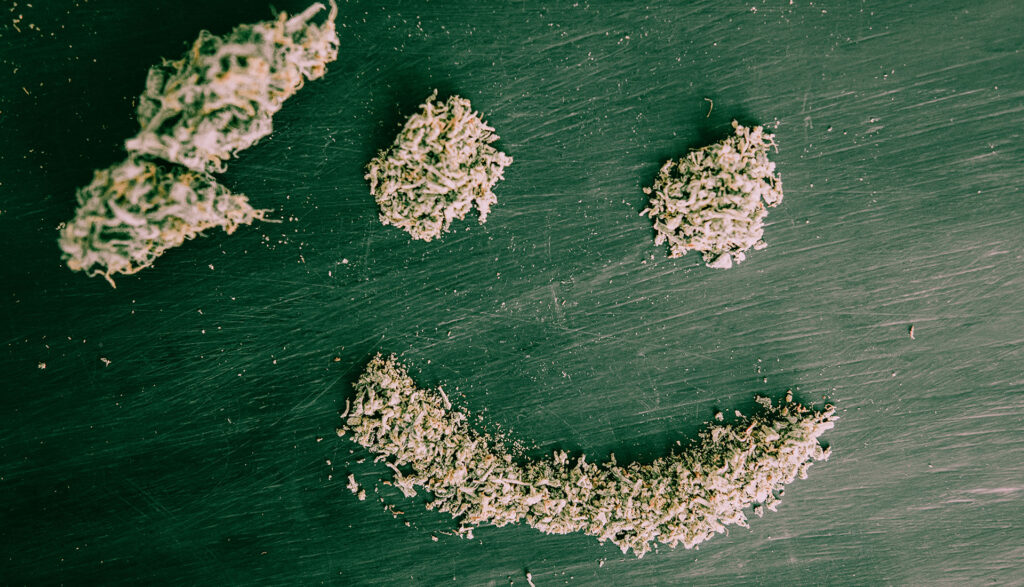 A smiley face shaped from cannabis flower on a green table background.